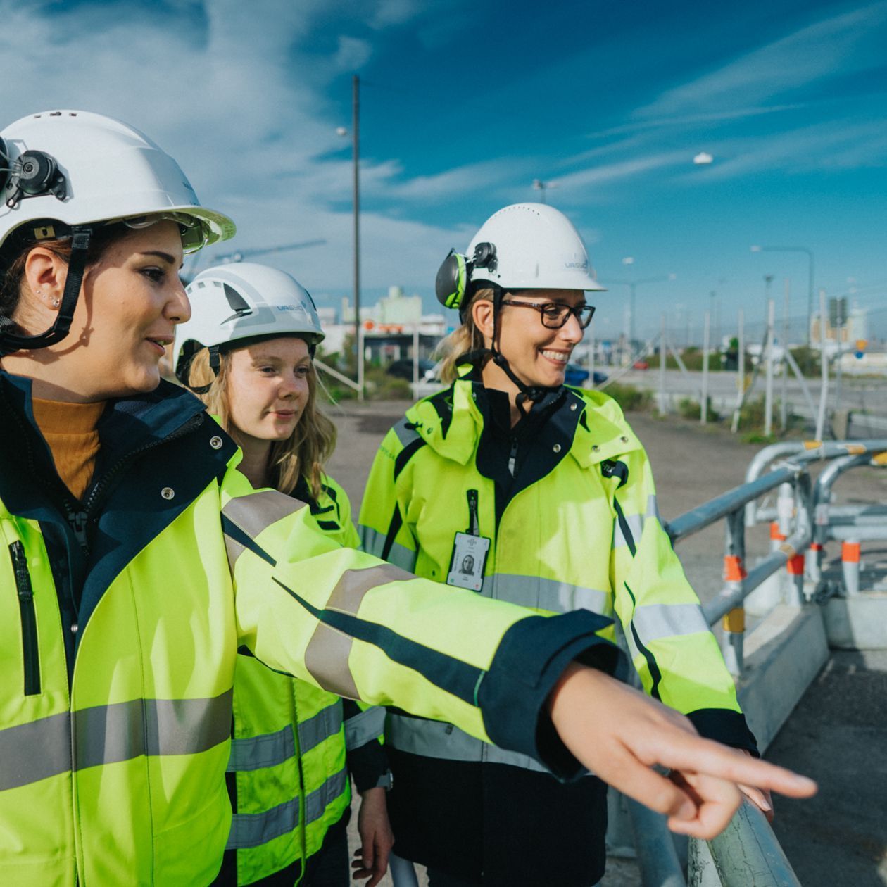 Tre personer i skyddsutrustning vid en industriell anläggning, där en person pekar framåt.