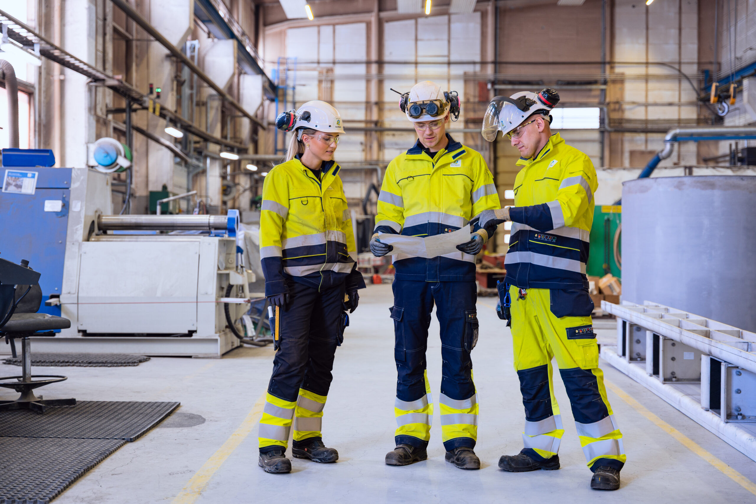 Tre industrimedarbetare i skyddsutrustning granskar en ritning i en industrimiljö